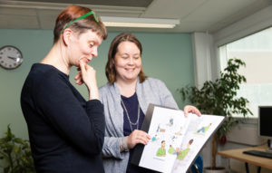 Sari Kivimäki ja Johanna Eskelinen selaavat kuvakommunikaatiokansiota. Kuvakommunikaatiota käytetään muun muassa työn ohjeistukseen ja perehdyttämiseen.