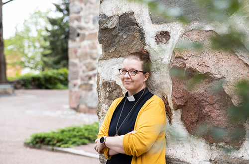 Lohjan seurakunnan pappi Pirkko Järvinen nojaa kivikirkon seinään. Papinkaavun päällä hänellä on iloisen keltainen villatakki.